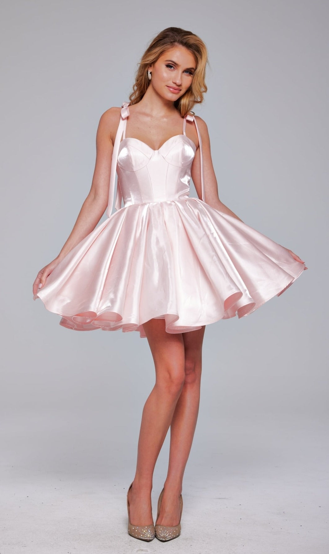 A woman models the Jovani Shoulder-Tie Short Pleated Hoco Dress 42342 in light pink with a flared skirt and sweetheart neckline, paired with beige heels against a plain gray background.